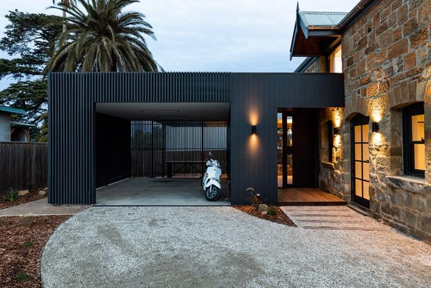 Exterior view of a contemporary addition to a stone building, featuring a dark ribbed-cladding carport or covered entry, exposed concrete paving, timber-lined entrance, and warm wall lighting. The original stone facade has arched openings and black-framed glazing, while a scooter is parked beneath the covered structure.