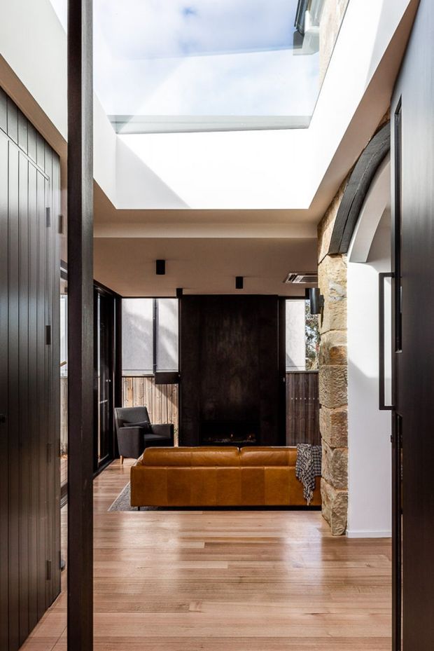 A refined contemporary living room viewed through a dark-framed entry, featuring warm timber flooring, a cognac leather sofa, dark timber wall panels, black-framed glazing, and a substantial exposed natural-stone arch and wall. A large skylight brings strong daylight into the room, creating a dramatic contrast between the bright ceiling and darker furnishings.