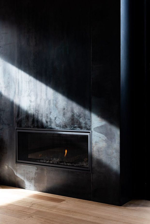 A dramatic, dark interior detail featuring a built-in linear gas fireplace set into a textured charcoal-black wall surround. Strong diagonal sunlight falls across the wall and fireplace, contrasting with the warm light timber floor. The composition is minimalist and architectural, with no visible furniture or room connection.