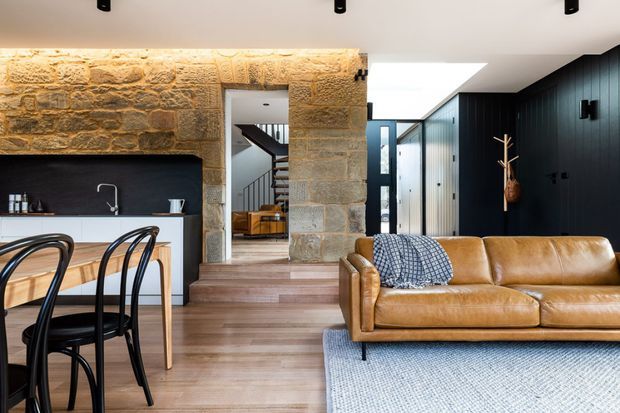 Open-plan interior combining a compact kitchen and dining area with a living room. A rough natural-stone feature wall and warm timber flooring contrast with dark vertical wall cladding and a bright white ceiling. The space includes a timber island or dining table with black bentwood chairs, a tan leather sofa, patterned cushions, and a pale textured rug. A stairway and entry corridor are visible beyond the stone opening.