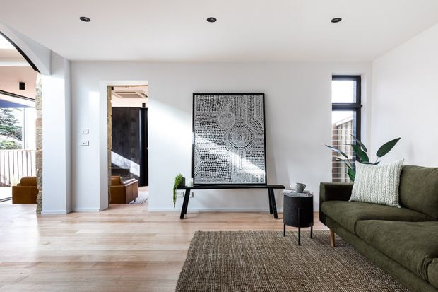 Bright contemporary living room with pale timber flooring, white walls and ceiling, recessed downlights, and a large olive-green sofa. A woven neutral rug, black side table, console, framed artwork, and indoor plant furnish the space. An open doorway leads to another interior area, while glazing at the left provides an outdoor glimpse.