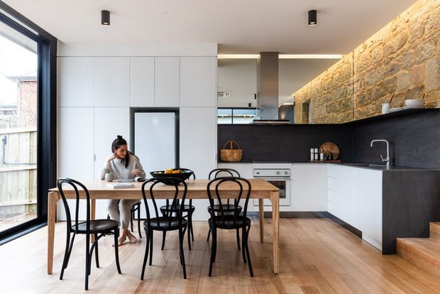 A bright, contemporary open-plan kitchen and dining area with a timber dining table and black bentwood-style chairs in the foreground. The kitchen features white flat-panel cabinetry, a dark benchtop and splashback, integrated appliances, a stainless-steel tap, and a prominent exposed rough-stone feature wall extending behind the cabinetry. Full-height black-framed glazing opens the room to daylight, while timber flooring and a small set of timber steps add warmth and visual continuity.