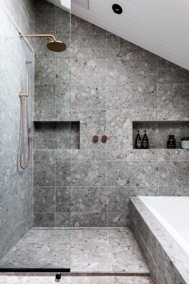 A modern bathroom featuring a walk-in shower clad floor-to-ceiling in large grey natural-stone tiles. The shower includes a slim copper-toned rain shower and handheld fixture, recessed wall niches with toiletries, and a partial freestanding or built-in white bathtub at right. The ceiling has white paneling and a recessed black downlight. The palette is predominantly cool grey stone with warm metallic accents.