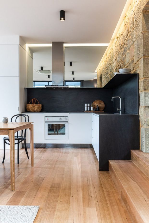 A contemporary open-plan kitchen and dining area with white flat-panel cabinetry, a dark charcoal island and splashback, integrated oven, stainless-steel sink, timber dining table and black chairs. Warm timber flooring continues up the adjacent steps, while a prominent rough natural-stone wall provides texture. Recessed linear lighting and black cylindrical downlights create a clean, modern fit-out.
