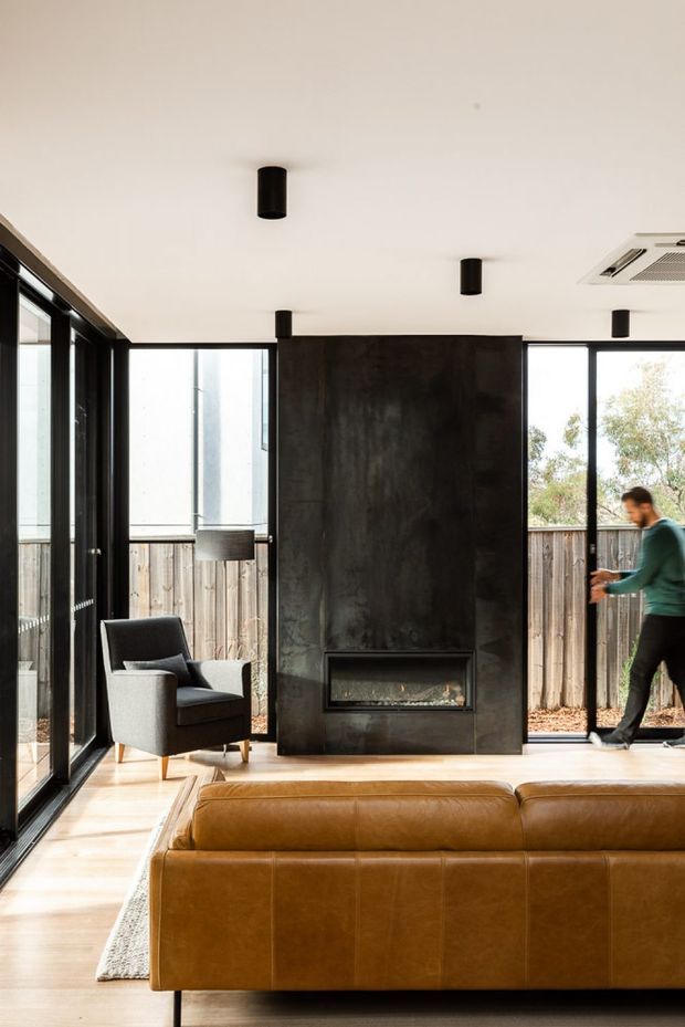 Bright contemporary living room with floor-to-ceiling black-framed glazing, a large dark stone or concrete fireplace feature wall with integrated gas fire, pale timber flooring, and a caramel leather sofa facing the fireplace. A grey upholstered armchair sits beside the glazing, with a neutral rug and minimalist black cylindrical ceiling lights. The room opens visually to a fenced outdoor area.