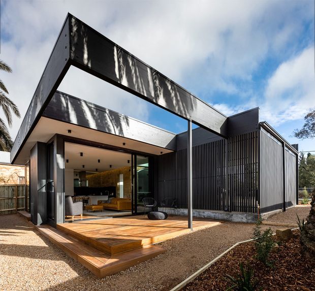 Contemporary single-storey house viewed from the landscaped exterior, featuring a dramatic projecting flat-roof frame, dark metal cladding and vertical screening. Large glazed openings connect the interior living area to a raised timber deck, with a visible kitchen and lounge beyond. Gravel landscaping and established planting surround the home.