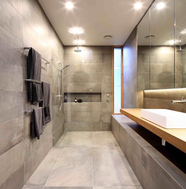 A contemporary bathroom with a spacious walk-in shower, large-format grey stone-look tiles on the walls and floor, a floating timber vanity with a white vessel basin, and a large mirrored wall. Recessed ceiling lights and concealed linear lighting create a clean, refined atmosphere.