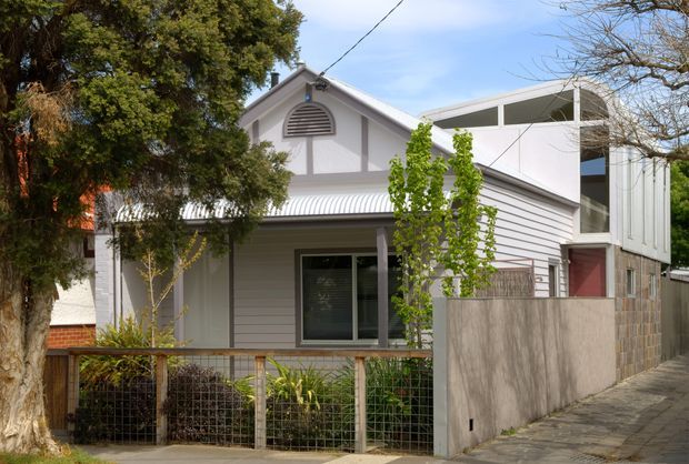 Street-facing view of a compact residential house with a white weatherboard facade, gabled front elevation, corrugated metal awning, and a contemporary side extension. The home features a small planted front garden behind a wire-and-timber fence, large windows, and a paved pedestrian approach. Mature trees partially frame the property.