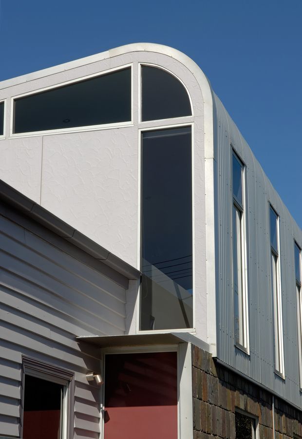 Close-up exterior view of a contemporary multi-storey building facade under a clear blue sky. The composition highlights a tall glazed entry or stairwell volume with a distinctive arched upper window, white rendered framing, grey-blue corrugated metal cladding, and contrasting horizontal weatherboard cladding. A dark red recessed doorway and lower masonry wall are visible at ground level.