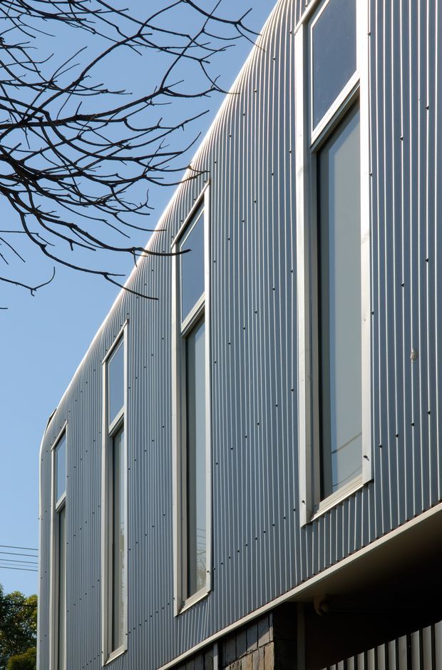 Close-up exterior architectural view of a contemporary building facade clad in vertically ribbed grey metal panels. Tall, narrow windows are arranged between the corrugated cladding bays, with a projecting lower edge and partially visible masonry base. Leafless tree branches frame the upper left against a clear blue sky.