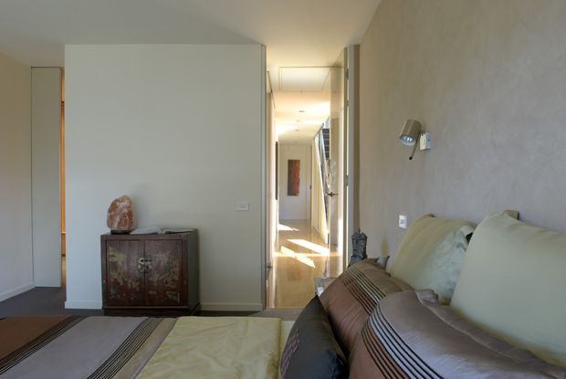 A narrow, softly lit contemporary interior viewed from a living area, with a large upholstered sofa in the foreground, a low decorative cabinet against a partition wall, and a long hallway extending toward the back of the home. Neutral plastered walls, recessed ceiling lighting, timber-toned flooring, and strong sunlight at the end of the corridor create a calm, understated atmosphere.