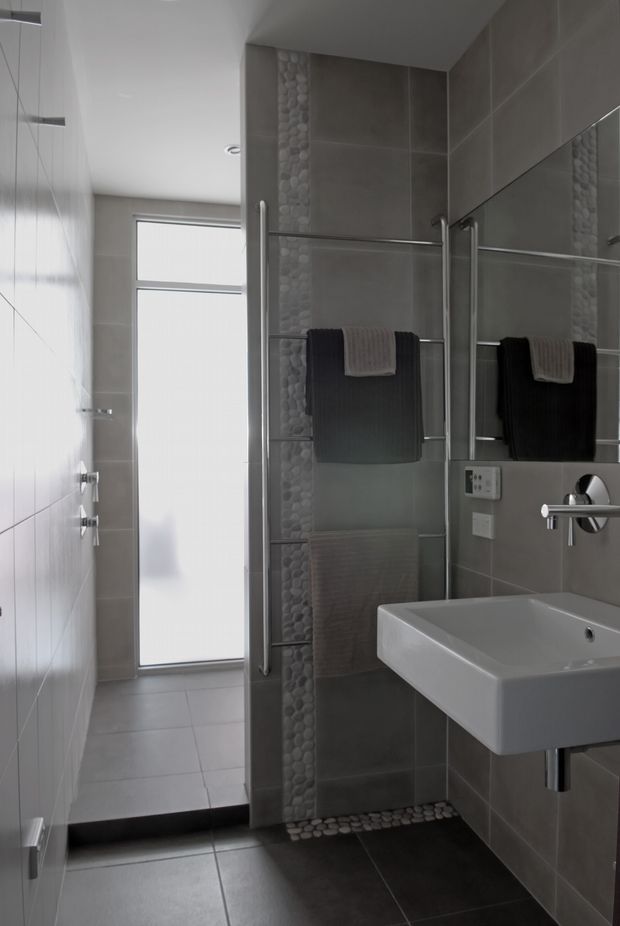 A compact contemporary bathroom with a wall-mounted white rectangular basin, frameless glass shower enclosure, large grey wall and floor tiles, and a vertical pebble-stone feature strip. Chrome towel rails, tapware, and fittings contrast with the grey finishes. A frosted full-height window provides diffused natural light, while a large mirror reflects the basin and towel rails.