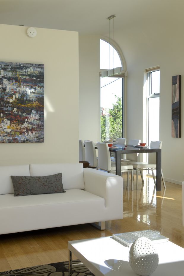 Bright open-plan interior with a white contemporary sofa in the foreground and a dining area behind it. The room has polished warm timber flooring, pale cream walls, tall windows including a prominent arched-top window, a dark dining table with white upholstered chairs, abstract artwork, and minimalist decor. Strong natural sunlight creates a clean, airy atmosphere.