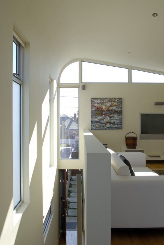 Bright contemporary living room viewed from a narrow elevated passage or stair landing, with dramatic clerestory and arched glazing, tall windows, white walls, a white sectional sofa, wall-mounted television, timber flooring, and strong sunlight. The split-level layout and external walkway or lower entry are visible below.