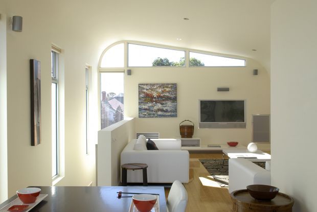 Bright open-plan living and dining area with a contemporary minimalist fit-out. The room features pale walls, timber flooring, a white sectional sofa, wall-mounted television, abstract artwork, recessed ceiling lights, and abundant natural light through tall windows and a distinctive curved clerestory window. A dining table occupies the foreground.