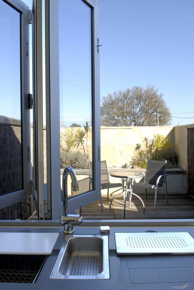 View from a compact kitchen through open white-framed glazed doors to a small timber deck or patio. The foreground shows a stainless-steel sink with draining board and chrome mixer tap set into a dark countertop, while the outdoor area contains a small round table, chairs, raised planter beds, and a rendered boundary wall.