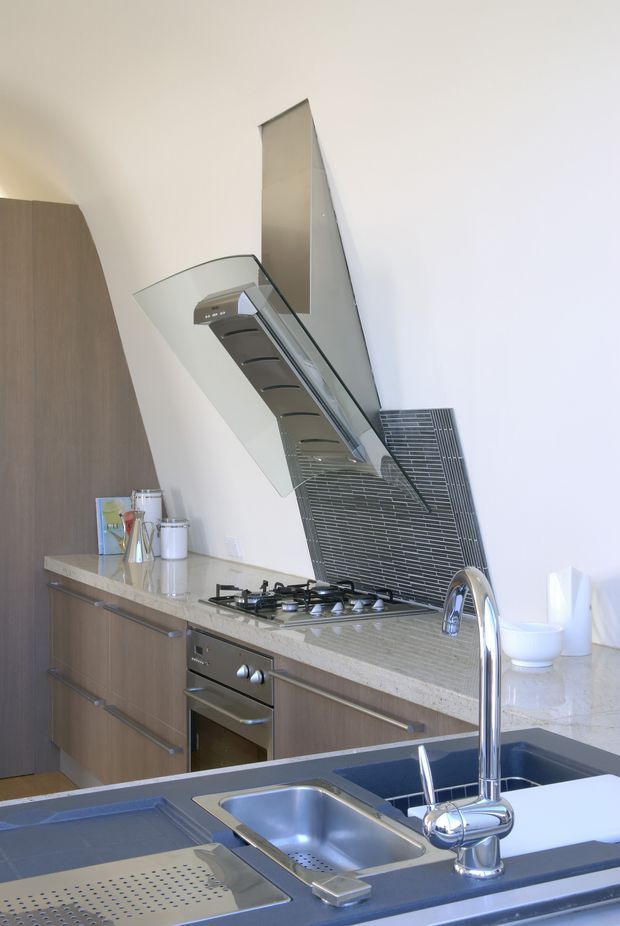 A contemporary residential kitchen viewed from the sink, with dark wood-grain cabinetry, a pale stone benchtop, stainless-steel appliances, gas cooktop, sculptural rangehood, and a narrow grey tiled splashback. The foreground features a double stainless-steel sink and chrome gooseneck tap, while a peninsula or island separates the sink from the cooking zone.