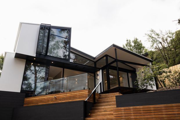 Exterior view of a contemporary multi-level house set into a wooded, sloping site. The building features crisp white rendered and dark cladding elements, extensive black-framed glazing, projecting angular roof planes, timber decking and stairs, and a transparent glass balustrade.