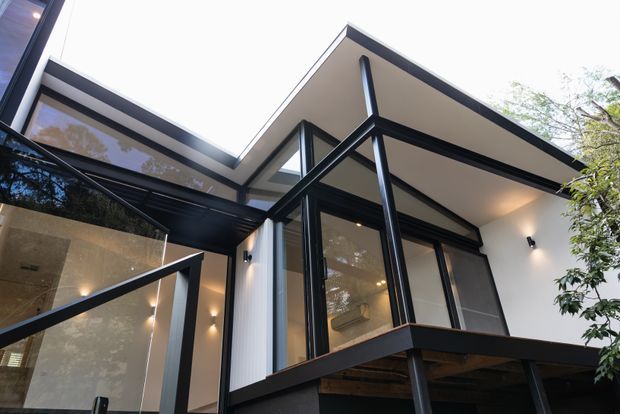 Upward-looking exterior view of a contemporary multi-level residence with sharply angled flat roof planes, extensive dark-framed glazing, projecting balconies, slim black structural posts, and illuminated white walls. Mature greenery surrounds the building.