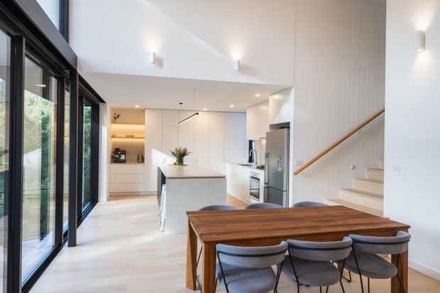 Bright open-plan kitchen and dining area with a long timber dining table and upholstered chairs in the foreground, a white minimalist kitchen with a central island behind, extensive black-framed glazing along the left side, and a staircase with a timber handrail at right. The space has a high sloping ceiling, pale timber flooring, white cabinetry and walls, and contemporary integrated lighting.