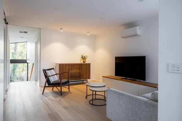 Bright contemporary living room with pale timber flooring, a grey upholstered sofa, black-framed lounge chair, nested round coffee tables, a timber sideboard, and a low white media console beneath a wall-mounted television. A glazed door opens to greenery, and a split-system air conditioner is mounted above the TV.
