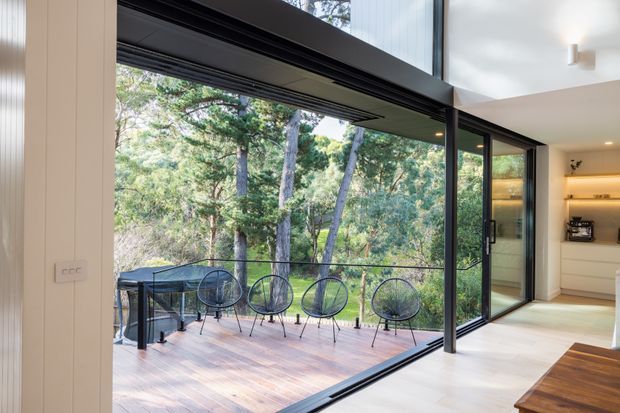 A bright contemporary interior opens through expansive black-framed sliding glass doors onto a timber deck surrounded by dense bushland. The room includes a minimalist kitchen or kitchenette with warm under-cabinet lighting, while the deck has a small outdoor dining setting and overlooks the trees. High ceilings and extensive glazing create a strong indoor–outdoor connection.