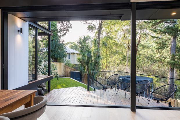 A bright contemporary interior opens through large black-framed sliding glass doors onto a raised timber deck and leafy garden. The space includes a timber dining table and curved chair in the foreground, with outdoor wire-frame seating on the deck, dark structural posts, pale timber flooring, and vertical wall cladding. The glazing provides an expansive connection to the surrounding trees and neighbouring garden.