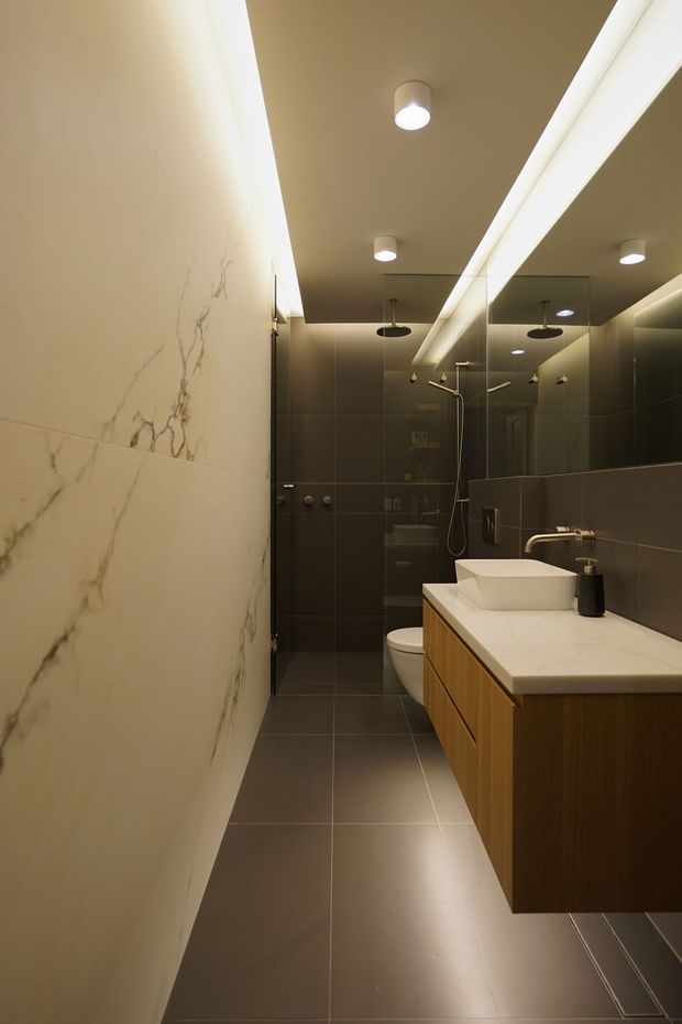 A narrow, contemporary bathroom with a walk-in glass shower at the rear, wall-hung toilet, floating timber vanity, white vessel basin and pale stone-look wall surface. Dark large-format floor and wall tiles create a warm, moody atmosphere, complemented by recessed ceiling lights and concealed LED lighting.