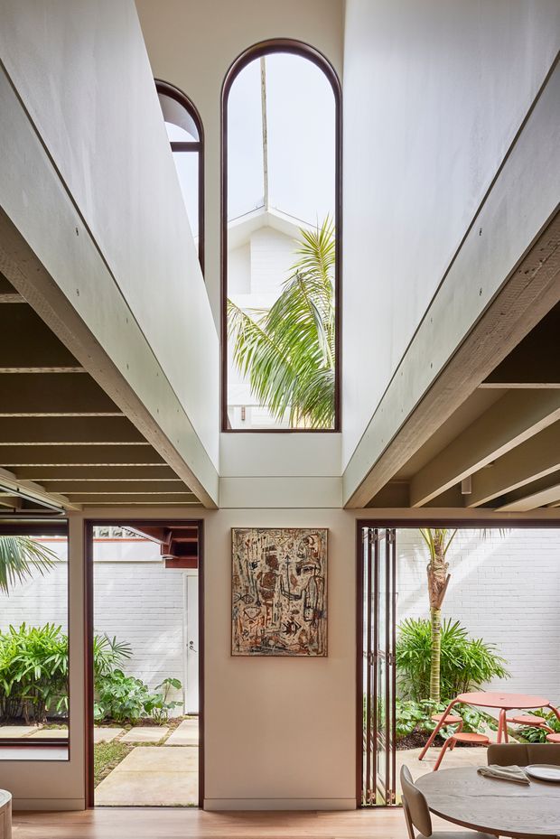 Interior view of a bright, double-height central circulation space or living area with exposed timber beam structures, white walls, and tall arched windows and doors framed in dark timber. The glazing opens toward a lush planted courtyard, while a dining table and chairs occupy the lower right. An artwork hangs on the central wall, and the space has a warm, contemporary architectural character with strong indoor-outdoor connection.