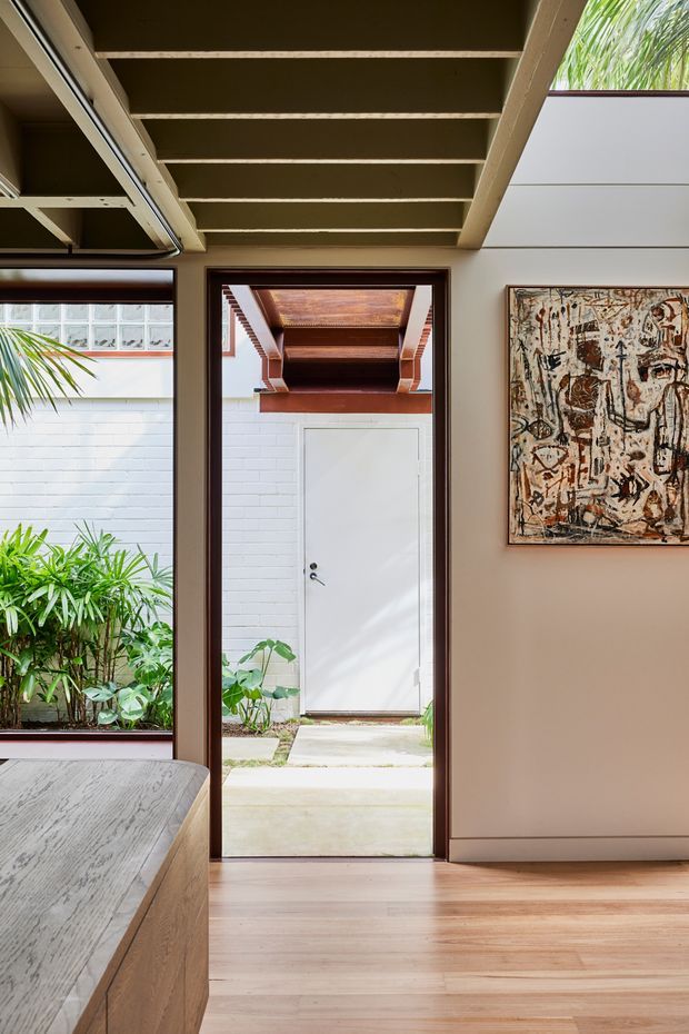 A bright interior space viewed toward an open doorway leading to a planted courtyard. The room has warm timber flooring, pale beige walls, dark timber door and window frames, and an exposed timber ceiling structure with deep beams. A built-in timber element occupies the lower left, while an abstract artwork hangs on the right wall. Through the doorway, a white-painted brick wall, a secondary white door, timber awning, paving, and lush tropical planting are visible. The composition has a refined mid-century residential character with strong indoor-outdoor connection.