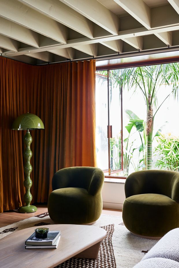 A richly styled living room with two rounded olive-green lounge chairs, a low light-wood coffee table, layered neutral rugs, and a sculptural green floor lamp. Full-height warm brown curtains frame a large glazed opening looking onto lush tropical planting. The ceiling has exposed painted structural beams, creating a striking contrast with the soft, curved furniture and warm natural light.