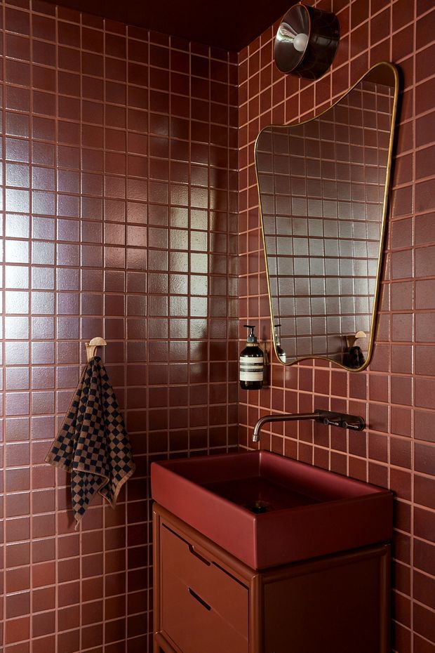 A compact, richly coloured bathroom corner with glossy burgundy-red square wall tiles, a matching red vessel sink and vanity, and dark metal plumbing fixtures. An irregularly shaped, gold-edged mirror hangs above the basin, accompanied by a wall-mounted soap dispenser and a patterned hand towel. The room has a moody, dramatic and cohesive vintage-inspired look.