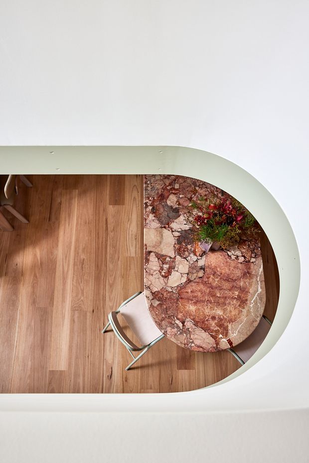 Top-down view of a refined interior seating area with warm timber flooring, a sculptural curved cream-coloured built-in wall or ceiling edge, and a rounded table made from richly veined reddish-brown natural stone. A light upholstered chair and floral arrangement complete the composition.