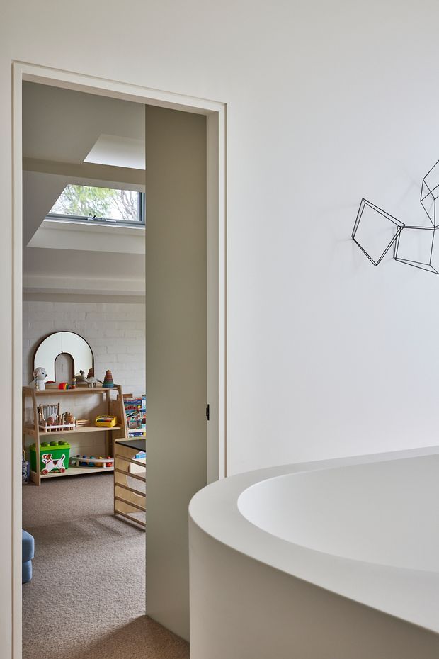 A calm, minimalist interior view from a bathroom or bathing area toward an adjoining children's room. The foreground is dominated by a large curved white freestanding bathtub with a smooth integrated surround. Through the open doorway, a playful children's room is visible with low timber shelving, toys, a carpeted floor, and a small arched mirror. The bathroom has white walls, neutral beige-grey tones, a skylight, and a partial white-painted brick wall. A simple black geometric line artwork appears on the wall beside the tub.