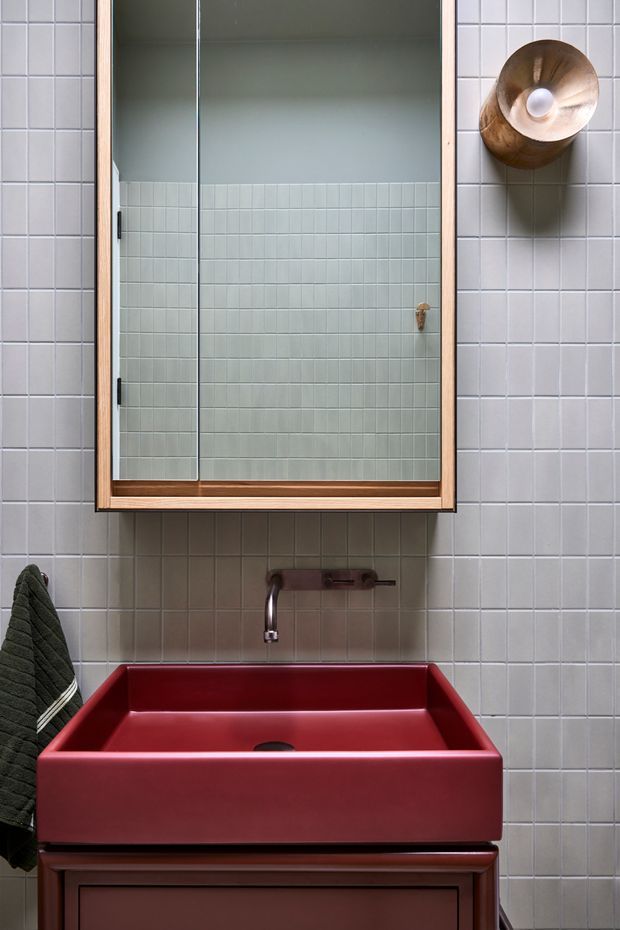A compact bathroom vanity scene with a deep red vessel-style rectangular basin on dark wood cabinetry, a wall-mounted metal tap, and a large timber-framed mirrored cabinet. Pale square wall tiles cover the room, with a dark towel and sculptural wall light adding contrast.