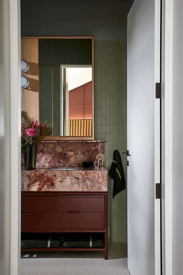A compact, richly styled bathroom viewed through an open white door. The room features a dark green tiled wall, a reddish-brown marble vanity splashback and benchtop with integrated basin, deep terracotta-brown timber cabinetry, a large framed mirror, and dark green accessories. The mirror reflects a brightly lit adjacent area with warm terracotta and timber detailing. The design has a moody, refined, colour-forward character.