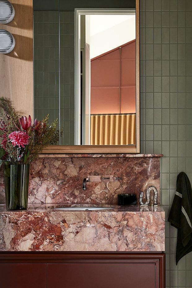 A richly detailed bathroom vanity vignette featuring deep green square wall tiles, a large timber-framed mirror, and a pink-red veined natural stone backsplash and benchtop with an integrated raised vanity surround. Dark reddish-brown timber cabinetry sits below, with a wall-mounted metal tap, black soap dispenser, sculptural chrome tapware, and a tall floral arrangement. The mirror reflects an exterior or adjoining architectural wall with warm terracotta and corrugated metal elements. The overall design is bold, refined, and richly layered.