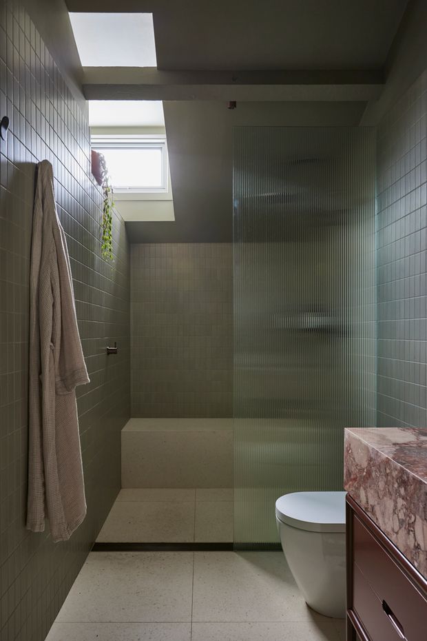A compact contemporary bathroom with full-height muted green square wall tiles, a terrazzo-like pale floor, and a walk-in shower separated by a ribbed translucent glass screen. A skylight and high window bring in natural light, while a marble vanity top with reddish veining and dark reddish-brown cabinetry occupies the foreground. A towel hangs on the tiled wall and a small trailing plant sits near the window.