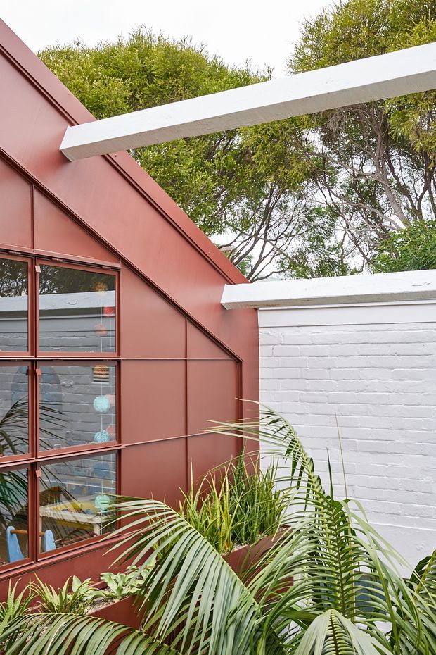 Close-up exterior view of a modern residential building courtyard or side elevation. The composition features a terracotta-red metal-clad gabled wall with dark-framed glazing, adjoining a white-painted brick wall with projecting white beams or roof edges. Lush palms and other planting fill the foreground, creating a warm contemporary architectural setting.