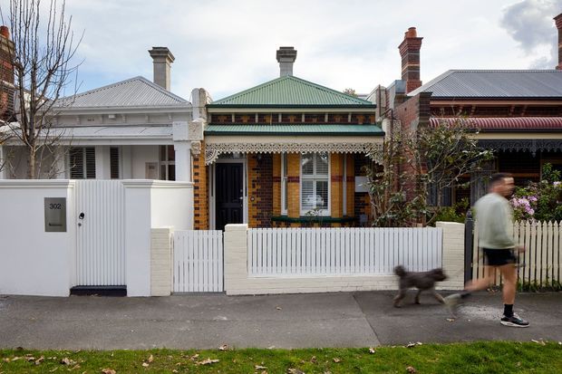 Street-facing view of a row of adjoining heritage terrace or townhouse facades. The central single-storey brick dwelling has a green corrugated metal hipped roof, decorative timber verandah detailing, brick chimneys, a white picket fence, and a small front gate. Neighbouring homes have similar period detailing and corrugated roofs. A pedestrian walking a dog is visible on the footpath.