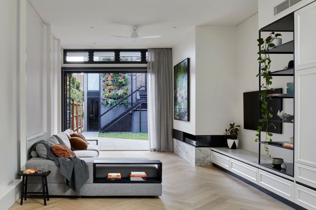 Bright contemporary living room with a grey sectional sofa, black-framed rear glazing and doors opening toward a small courtyard, pale herringbone timber flooring, white walls, and a built-in media wall with white cabinetry, black shelving, and marble-look cladding. Vertical curtains, indoor plants, and restrained furnishings create a clean, refined interior.