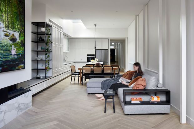 Bright open-plan living and dining area with a contemporary white kitchen along the left wall, integrated cabinetry, a dining table, and a grey sectional sofa with chaise in the foreground. The room features light herringbone timber flooring, white paneled walls, black open shelving, a marble-look fireplace surround or media unit, and a large artwork. Natural light enters through tall windows or glazed openings on the right, creating an airy, refined interior.