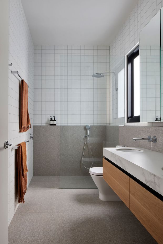 A contemporary bathroom with a walk-in shower, wall-hung timber vanity, white marble-look benchtop, wall-mounted basin tap, toilet, and large mirrored cabinet. The room is lined with small white square wall tiles, while grey speckled tiles or solid-surface panels cover the lower walls and floor. Black-framed glazing brings in natural light, and rust-coloured towels add warmth.