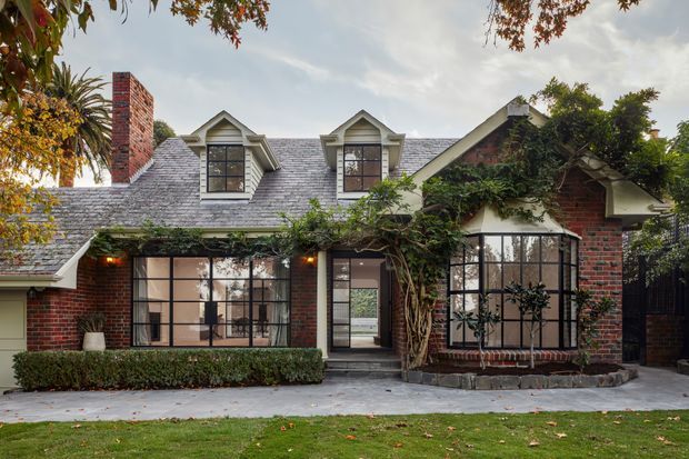 Front exterior view of a brick cottage-style residence with a steep pitched gabled roof, dormer windows, prominent brick chimney, and extensive black-framed glazing. Mature climbing greenery covers parts of the facade, with a paved entry path, manicured hedge, and lawn in the foreground.