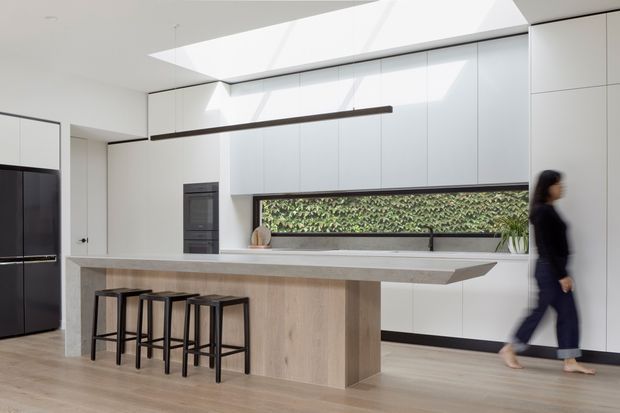 Bright, minimalist contemporary kitchen with a long pale stone island and integrated timber-toned base, four black stools, full-height white cabinetry, dark appliances, and a narrow planted window viewed beneath a large linear skylight. A slim dark pendant runs above the island, while pale timber flooring and restrained detailing create a clean, airy composition.