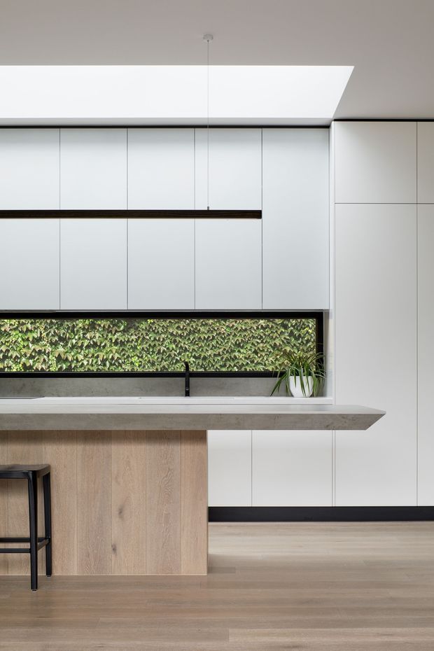 A minimalist contemporary kitchen with floor-to-ceiling white handleless cabinetry, a light timber island base, and a thick grey concrete-look benchtop with a projecting edge. A long horizontal window brings in greenery, while black detailing, a suspended linear pendant, and pale timber flooring add contrast.