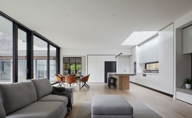 A bright, open-plan contemporary living, dining and kitchen interior. The space features a large grey sectional sofa and ottoman in the foreground, a dining area with sculptural timber-toned chairs, and a minimalist white kitchen with a timber island and dark bar stools. Full-height black-framed glazing lines the left side, while a large skylight brings additional natural light into the room. Light timber flooring, white cabinetry and walls, and restrained furnishings create a clean, airy atmosphere.