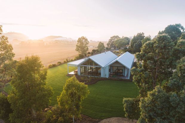 Aerial view of a contemporary single-storey residence set within a manicured rural landscape. The house has multiple intersecting pitched gable roofs, light-coloured exterior walls, extensive glazed sections, and a covered outdoor terrace. Mature trees surround the lawn, with open agricultural fields and rolling hills visible beyond in warm late-afternoon light.