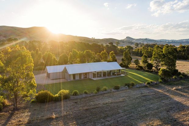 Aerial view of a long, low rural residence with pale metal pitched roofs, verandah-like covered outdoor areas, and light exterior walls. The home sits within broad lawns and mature trees, surrounded by cultivated fields and rolling hills in warm late-afternoon sunlight.