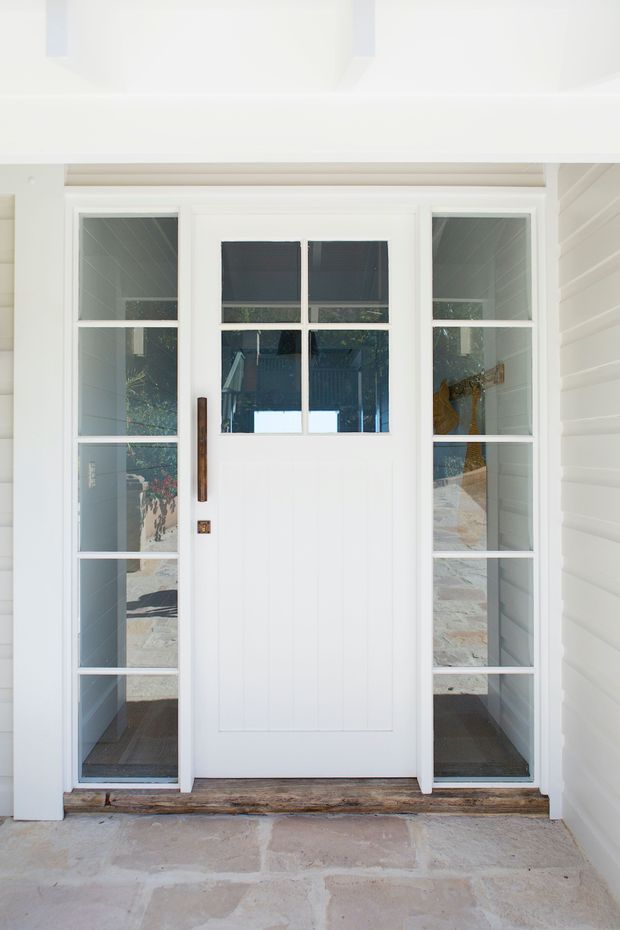 Front entry exterior featuring a white paneled timber door with divided-light glazing, narrow glazed sidelights, dark timber pull handle, and white horizontal weatherboard cladding. The doorway is set within a small covered porch with a pale stone-paved threshold.